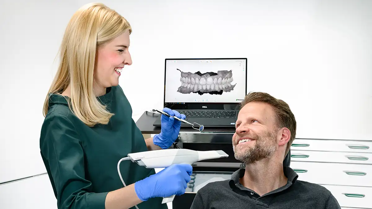 Zahnärztin hält Intraoralen Scanner und lächelt Patienten an. - im Hintergrund ein Bildschirm mit Scanergebnis