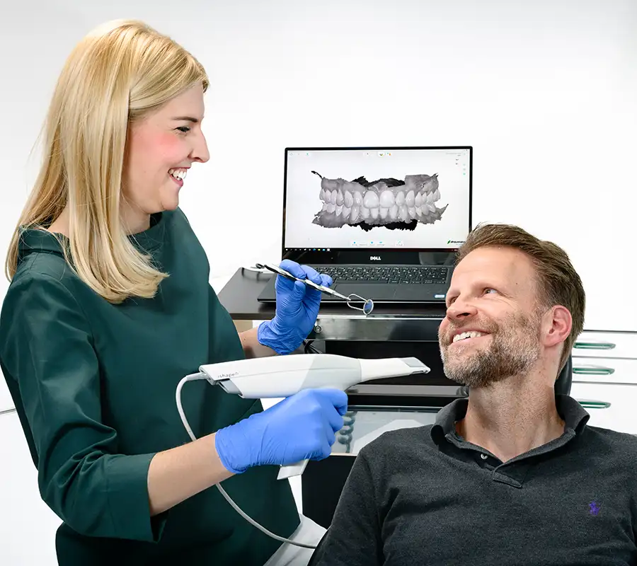 Zahnärztin hält Intraoralen Scanner und lächelt Patienten an. - im Hintergrund ein Bildschirm mit Scanergebnis