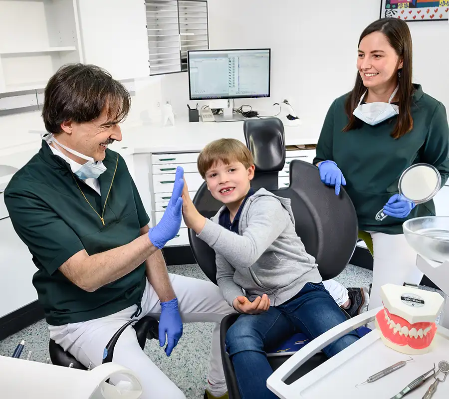 Zahnarzt klatscht Jungen im Behandlungsstuhl ab - im hintergrund lächelnde Helferin